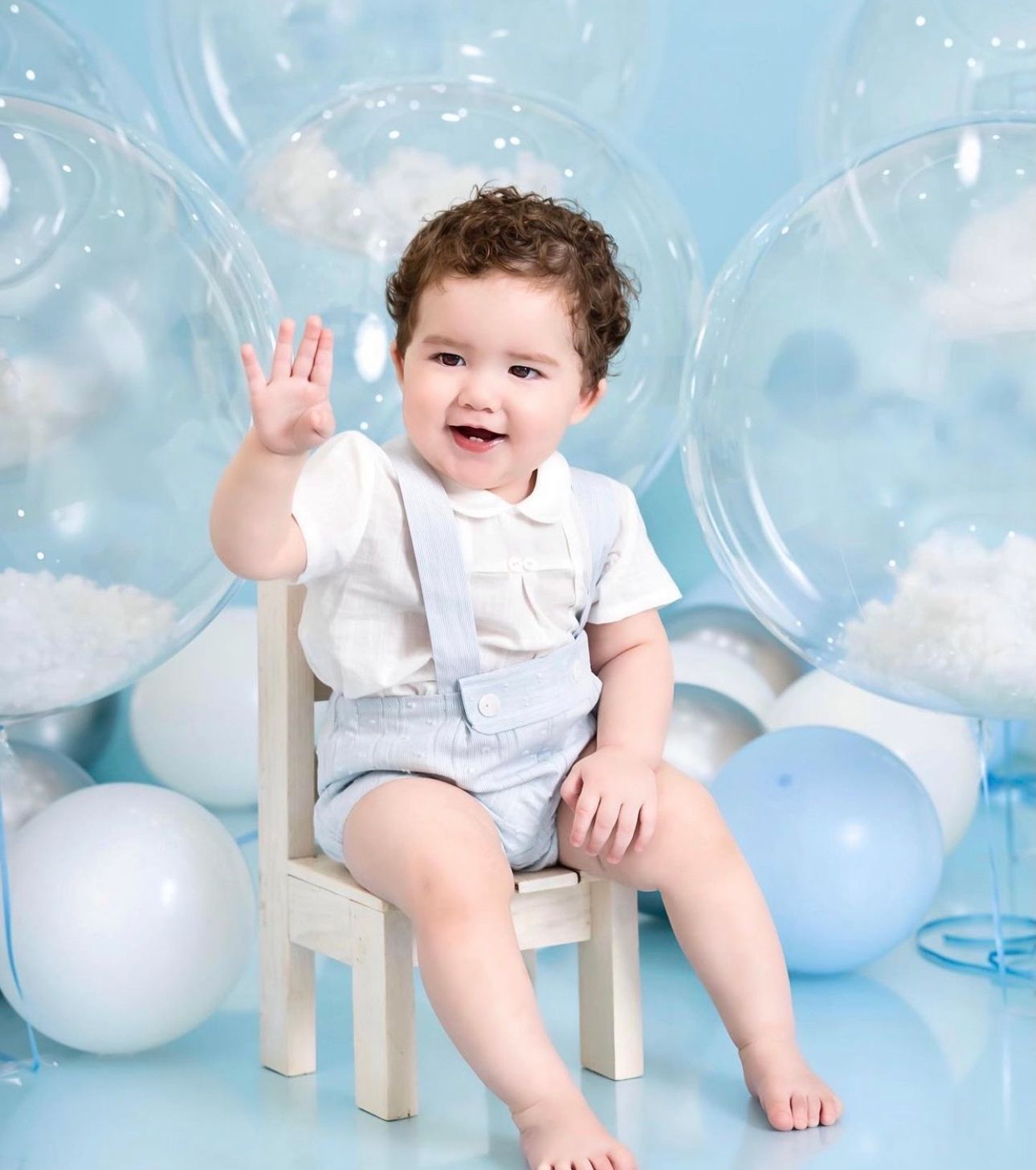 Baby in white outfit sits among light blue balloons and white decorations during a festive photo session.