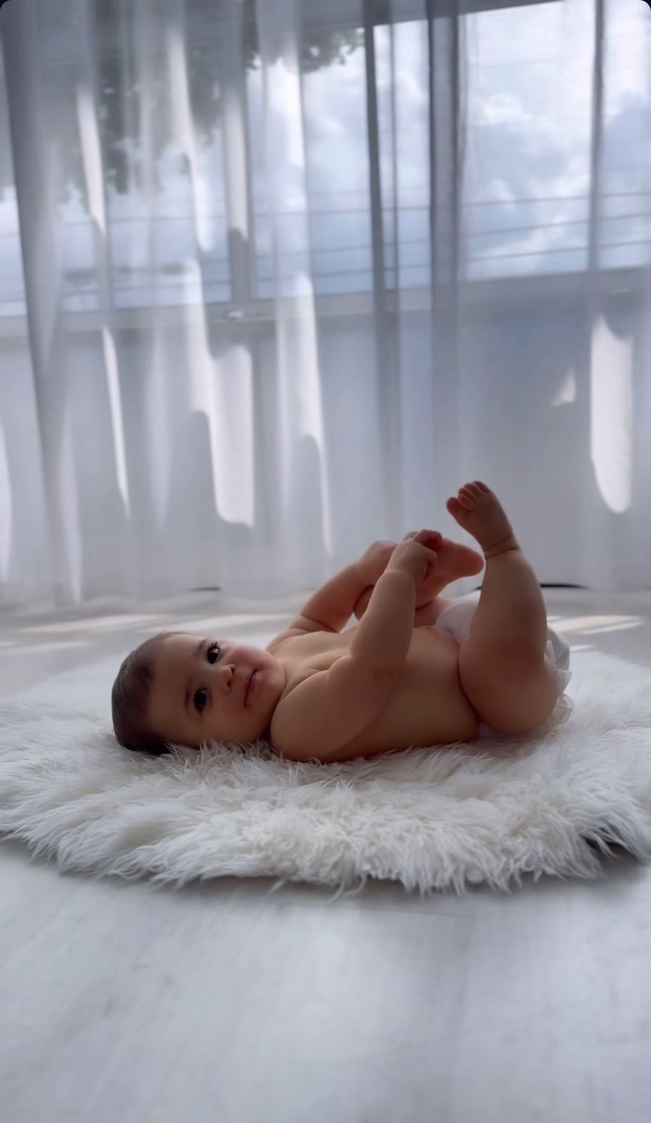Baby lying on white fluffy rug with soft window light creating peaceful atmosphere.