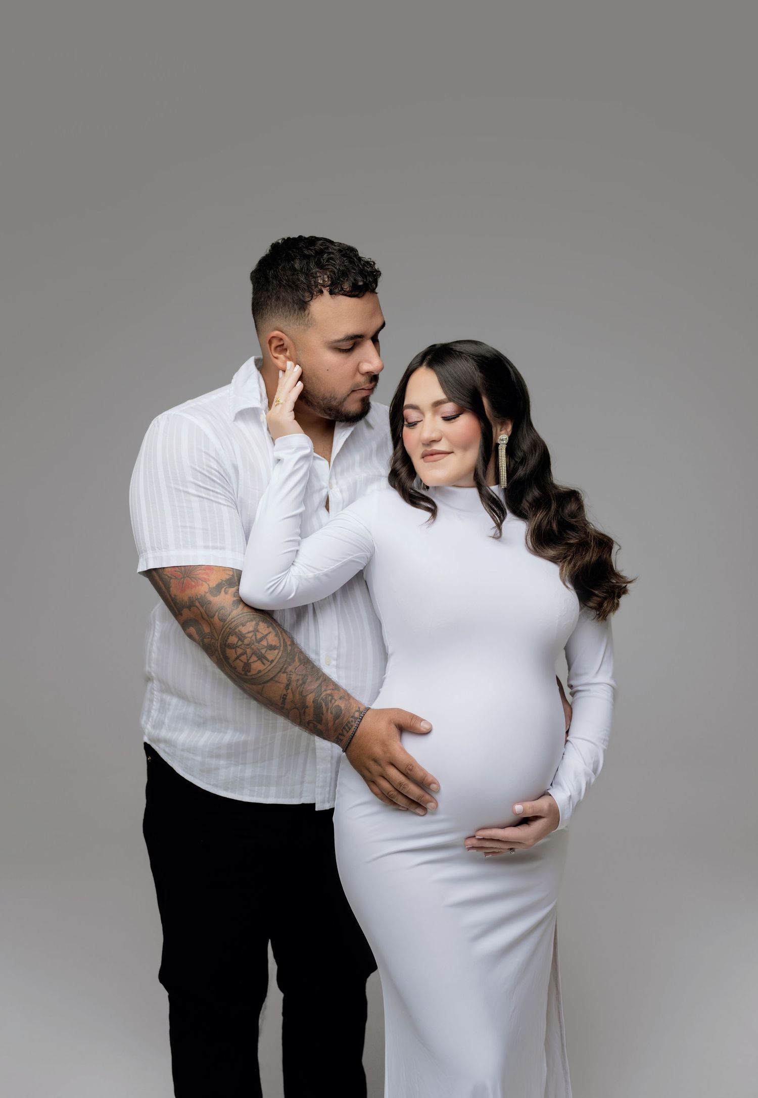 A couple in white clothing shares tender moments during a maternity photo shoot against a gray studio backdrop.