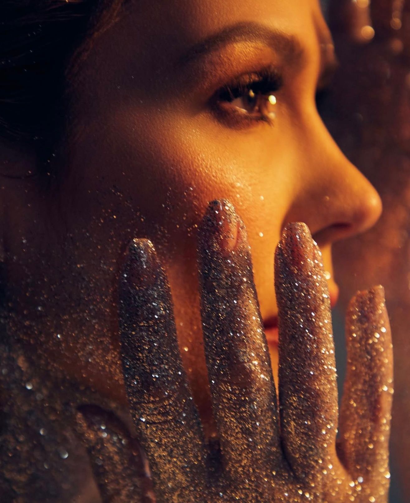 Close-up of a woman with glitter-covered hands pressed against her face, warm golden lighting highlighting her features.