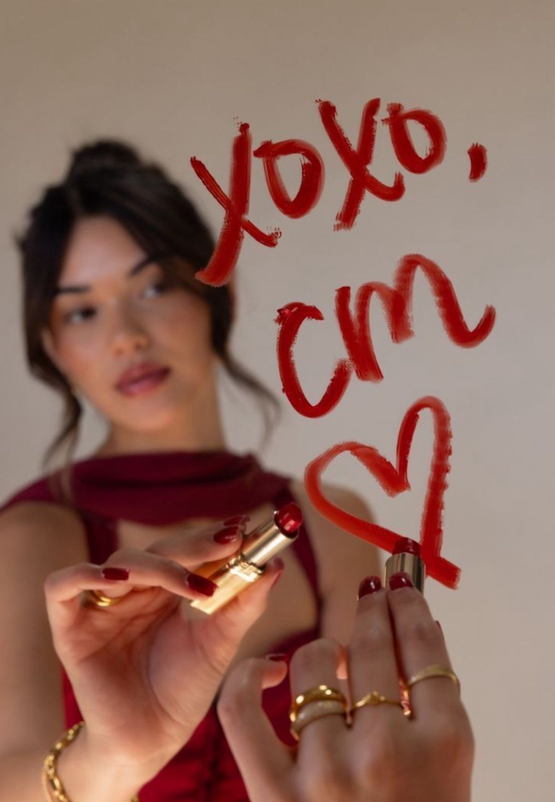 Woman in red dress writing XOXO, CM with red lipstick on glass, holding lipstick with gold rings.