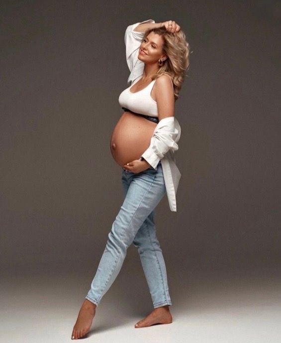 Pregnant woman in white tank top, open shirt, and light jeans posing for a maternity photo shoot.