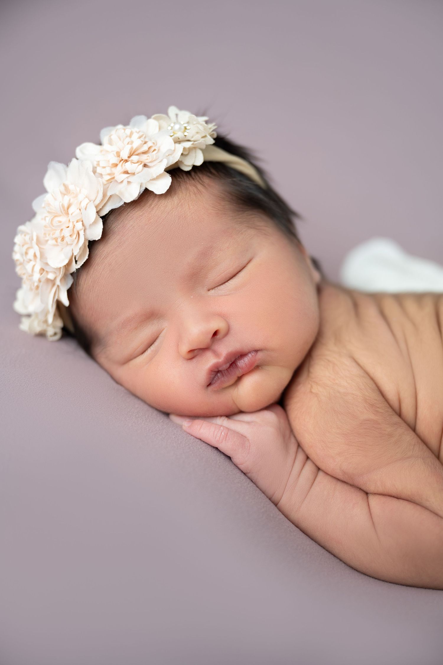 Sleeping newborn baby girl wearing a delicate white floral headband, resting peacefully on a soft purple background.