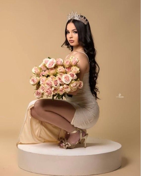Young woman in elegant cream dress and crystal crown holds pink roses bouquet, seated on white pedestal.