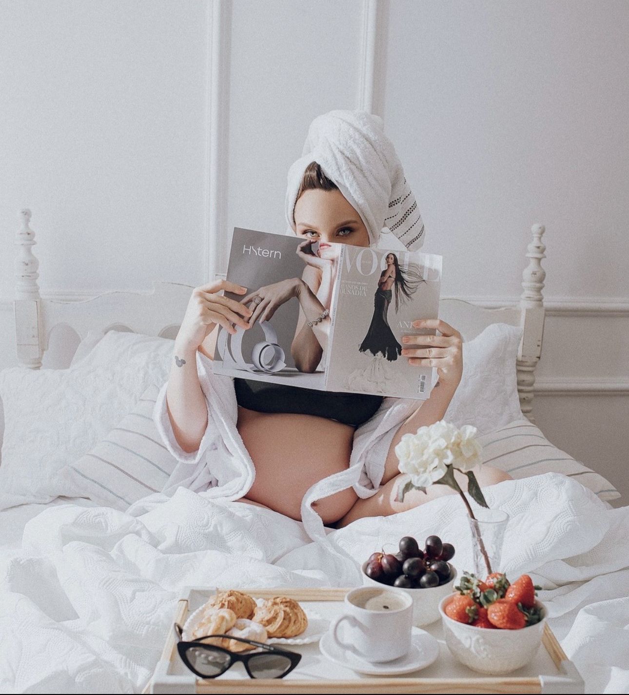 Pregnant woman in white robe and towel turban reads magazine in bed with breakfast tray of fruit and coffee.