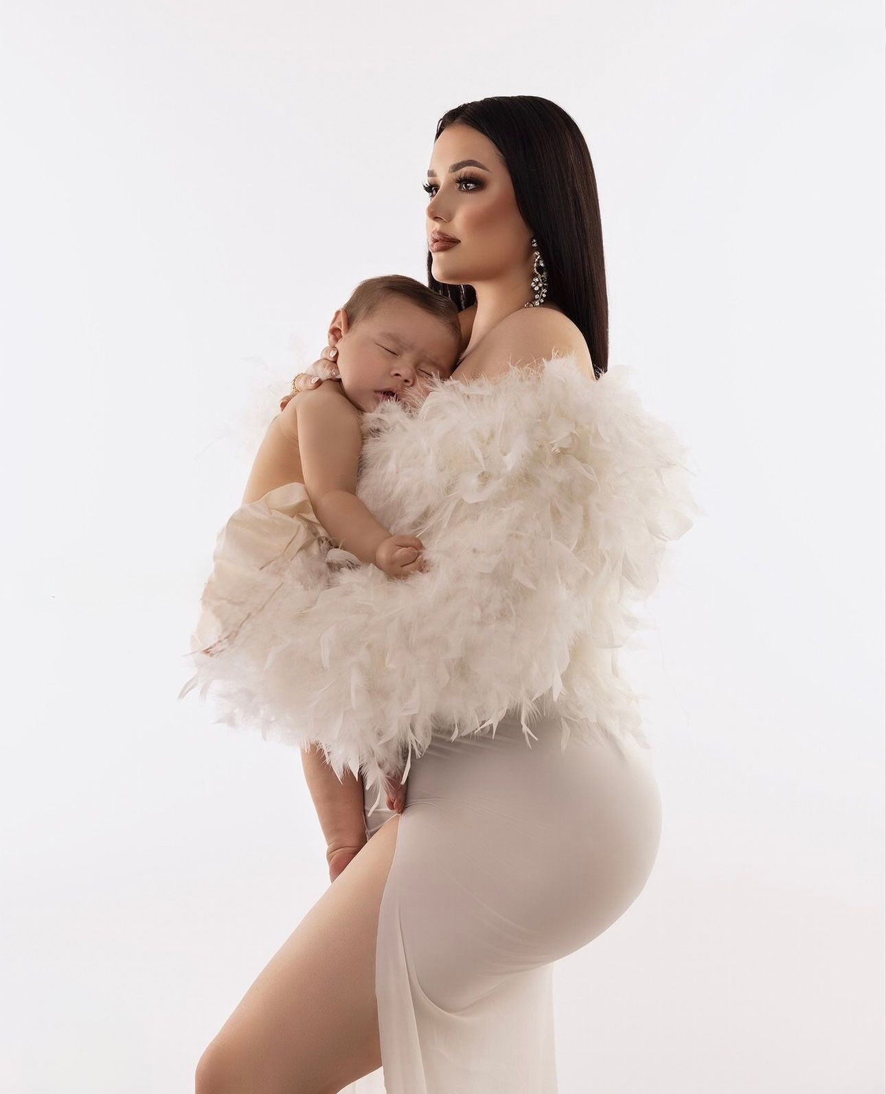 A woman in a white feathered wrap and fitted skirt holds a sleeping baby against a white background.
