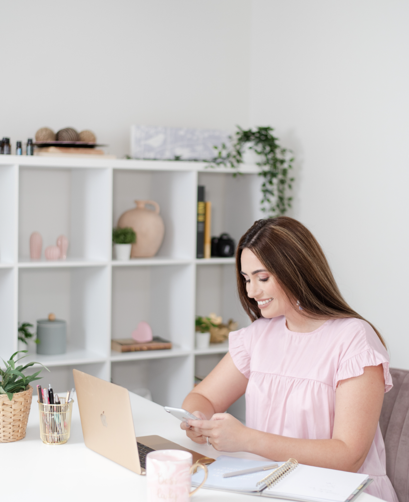Smiling woman in pink top using tablet at white desk with laptop, shelving unit, and plants in bright home office.