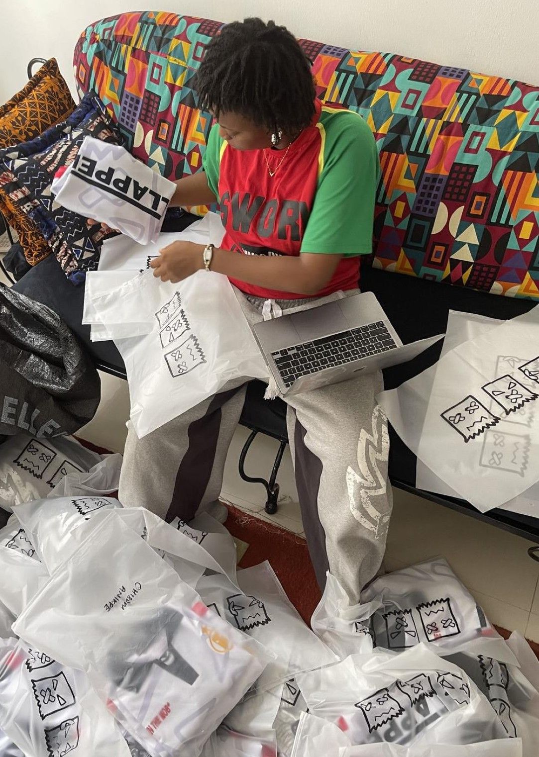 Person sorting branded shopping bags with a laptop, surrounded by merchandise orders on a colorful couch.