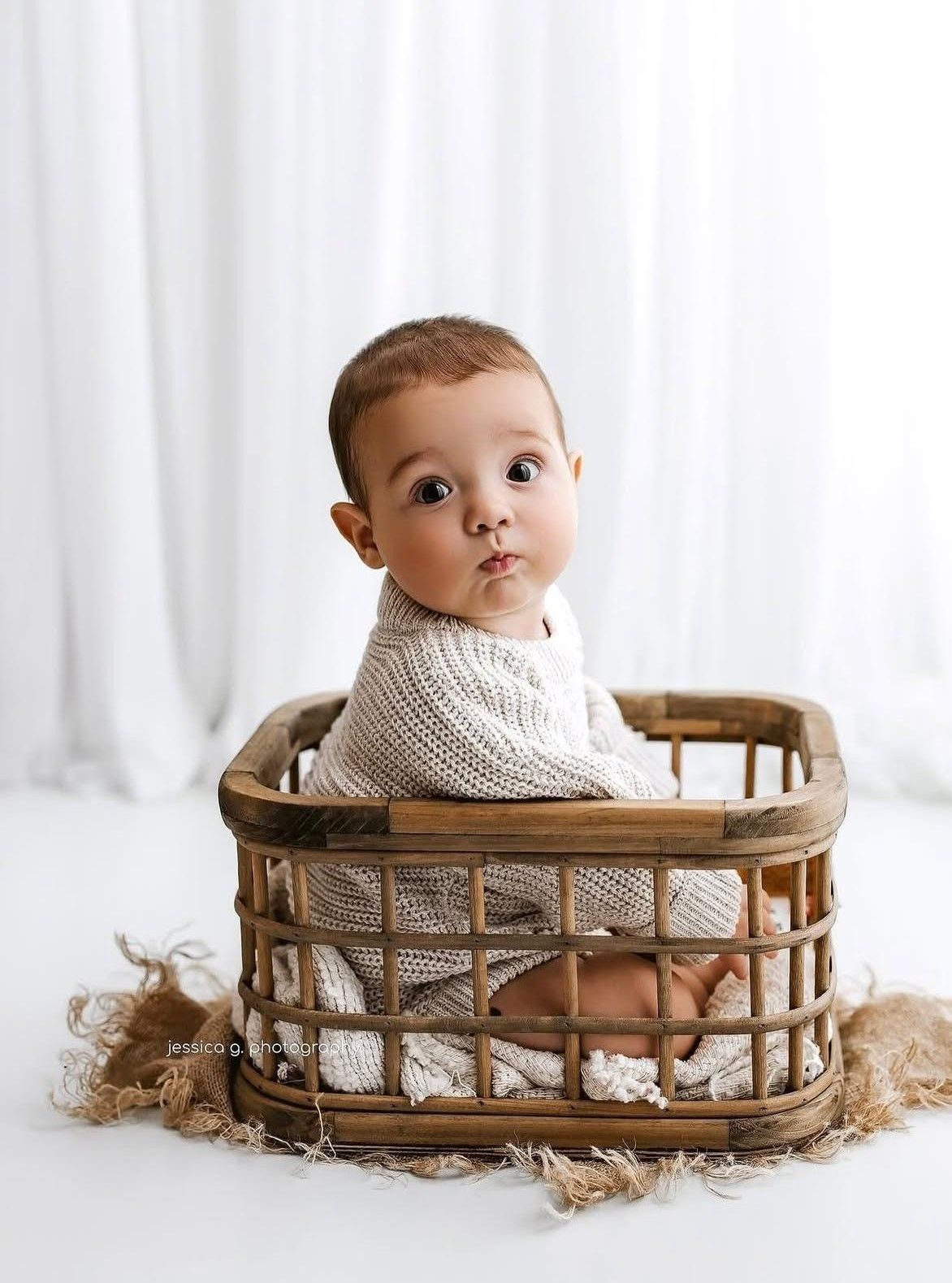 Adorable baby sitting upright in a rustic wicker basket, wearing a cream knit outfit, with a curious expression.