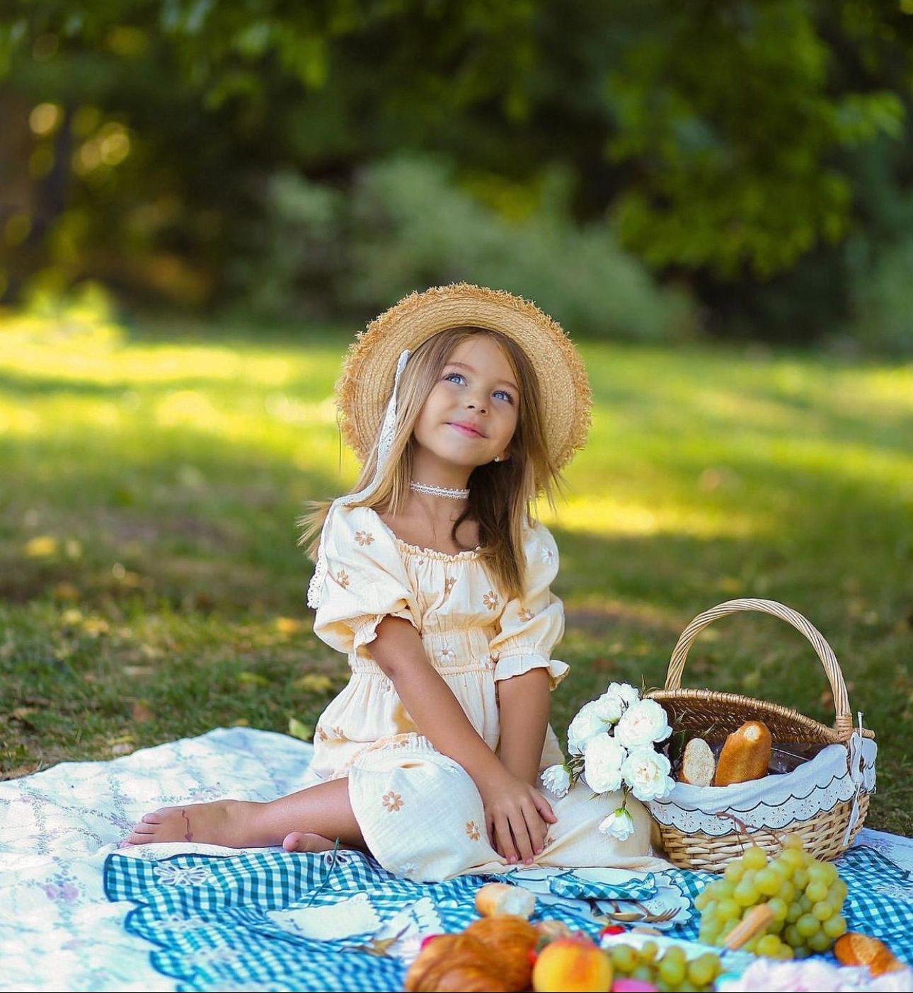 A young girl in a yellow dress and straw hat sits on a picnic blanket with a fruit basket in a sunny park.