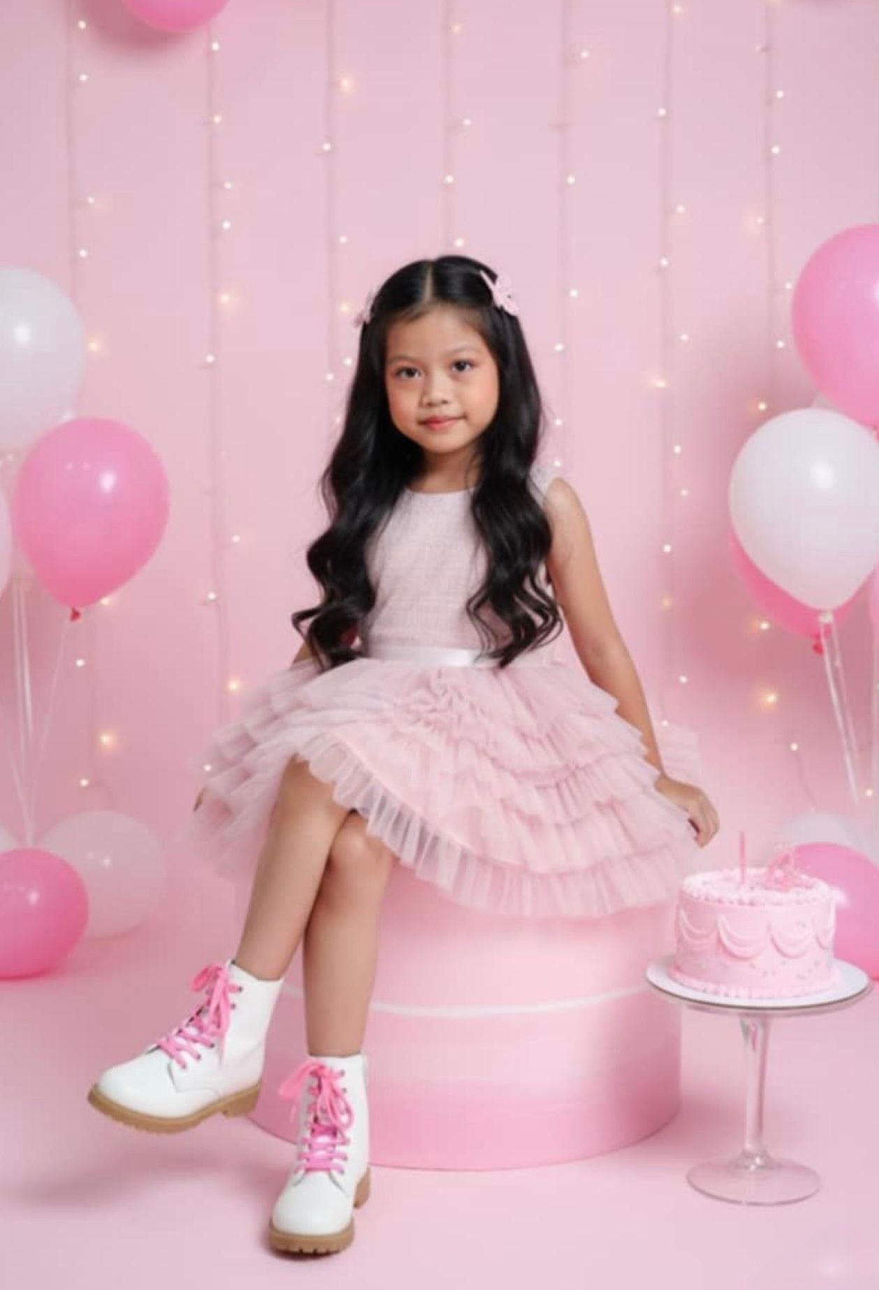 Young girl in pink tulle dress and white boots sits beside a pink birthday cake surrounded by pink balloons.