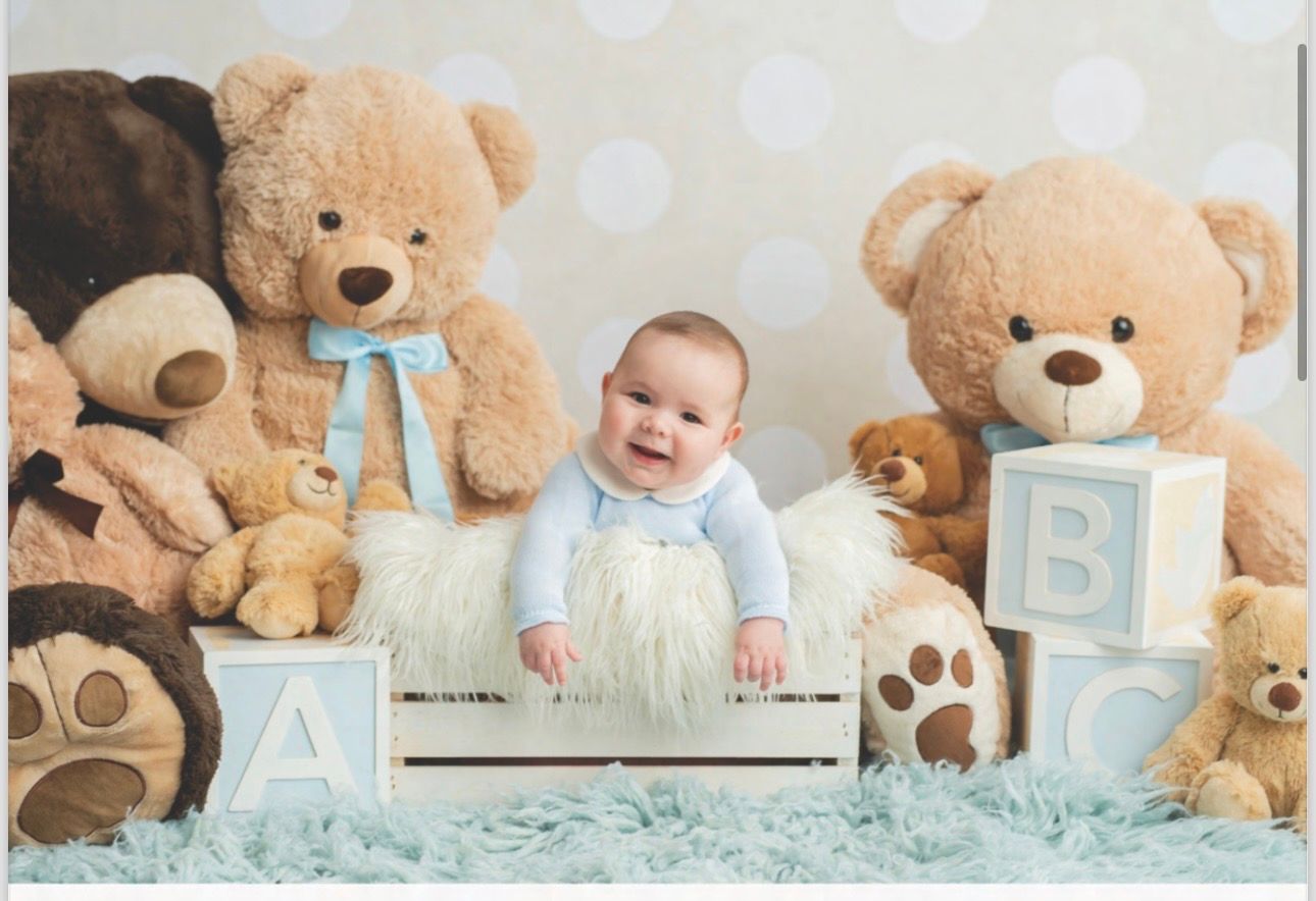 Smiling baby sits in white crate surrounded by large teddy bears and ABC blocks on blue fur rug.