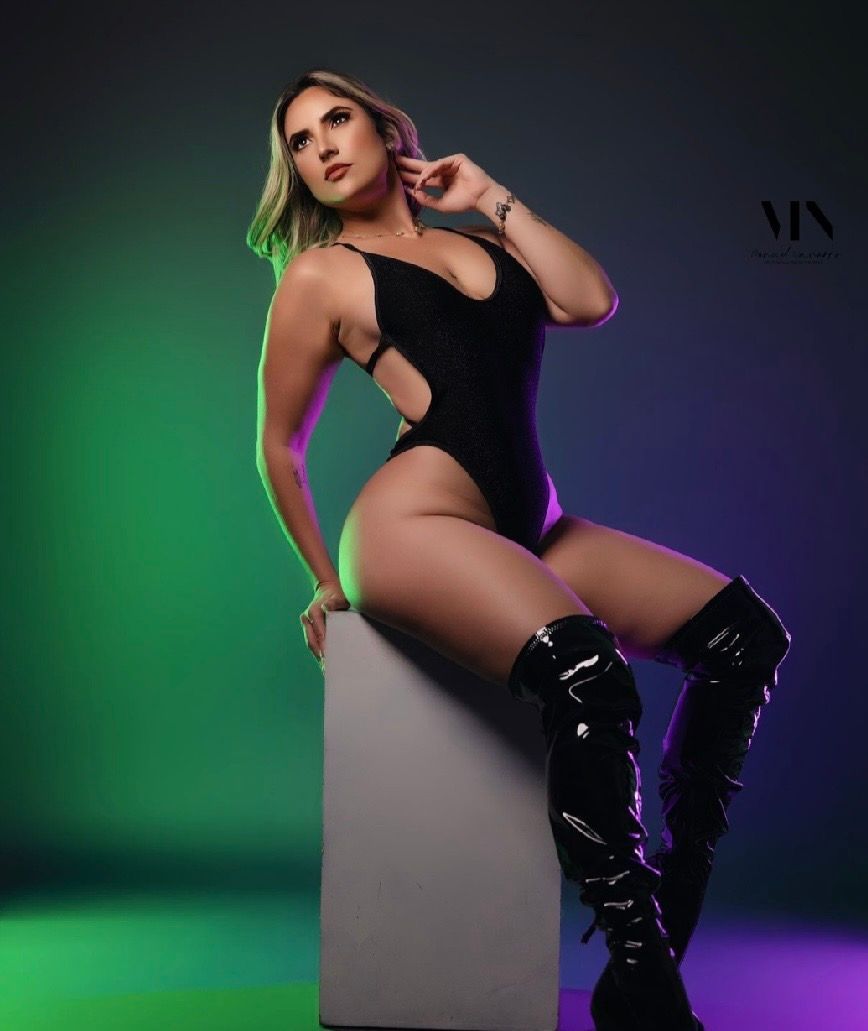 Woman in black bodysuit and thigh-high patent boots poses confidently against colorful green and purple studio backdrop.