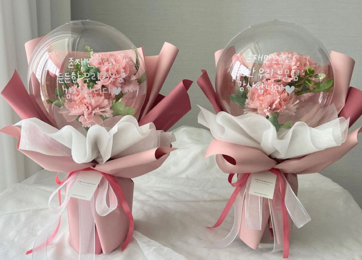 Two pink carnation bouquets in clear bubble balloons with pink ribbon wrapping on a white surface.