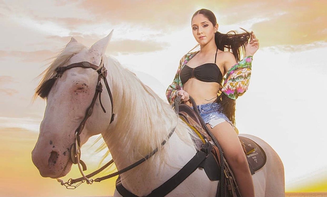 Young woman in bikini top and denim shorts riding a white horse at sunset with golden sky background.