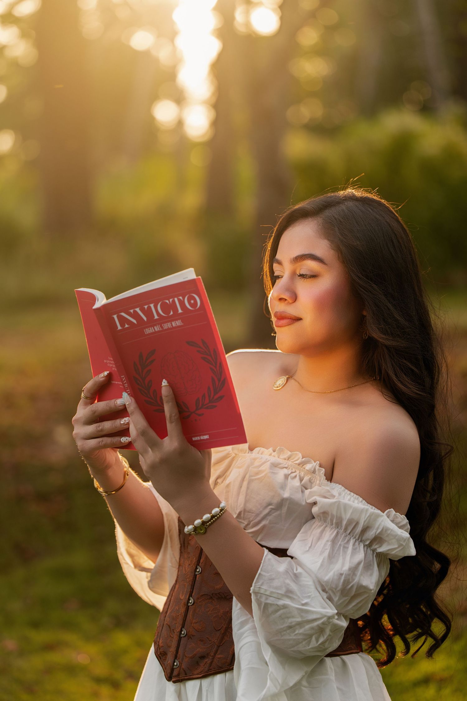 Young woman in white off-shoulder dress reads red Invicto book outdoors in golden sunlight forest setting.