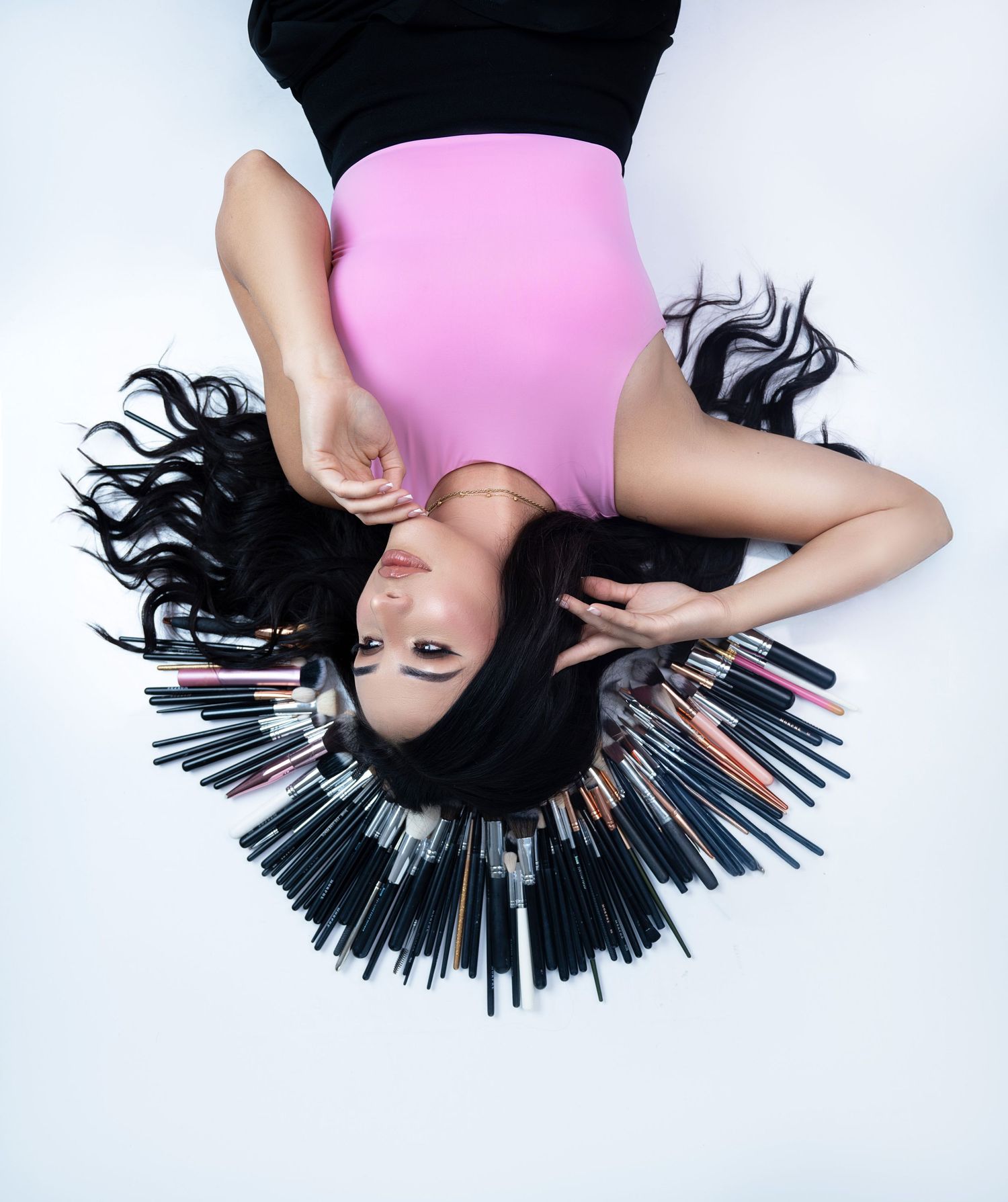Woman in pink bodysuit lying on white floor surrounded by makeup brushes arranged in a halo pattern around her dark hair.