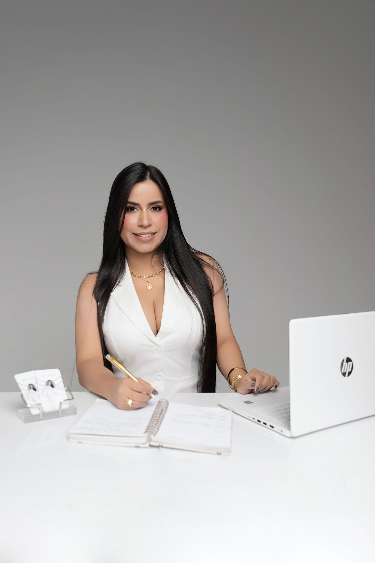 Smiling woman with long dark hair in a white blazer sits at a white desk writing in a planner beside an HP laptop.
