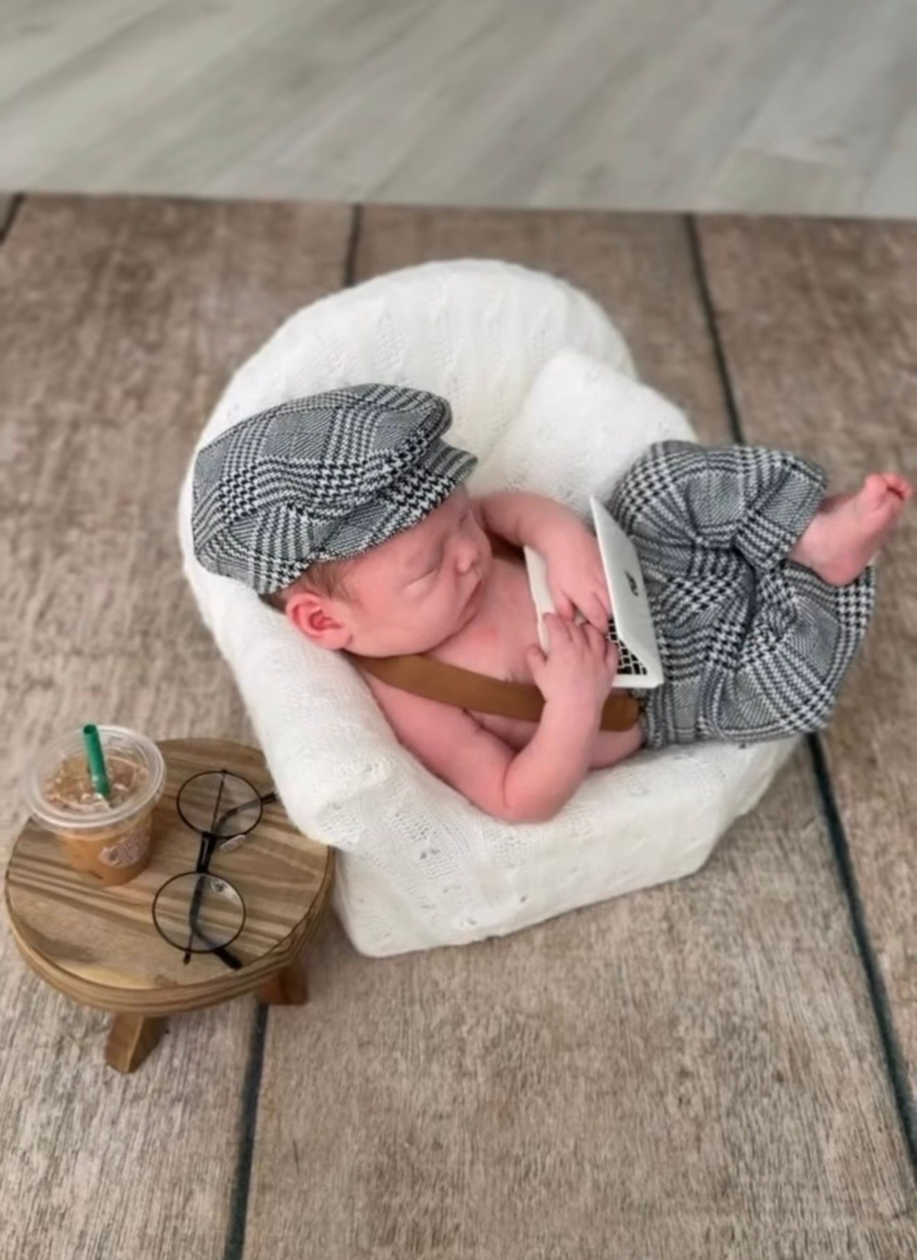 Adorable newborn baby dressed in plaid newsboy cap and suspenders, posed in fluffy white chair with tiny Starbucks cup and glasses.
