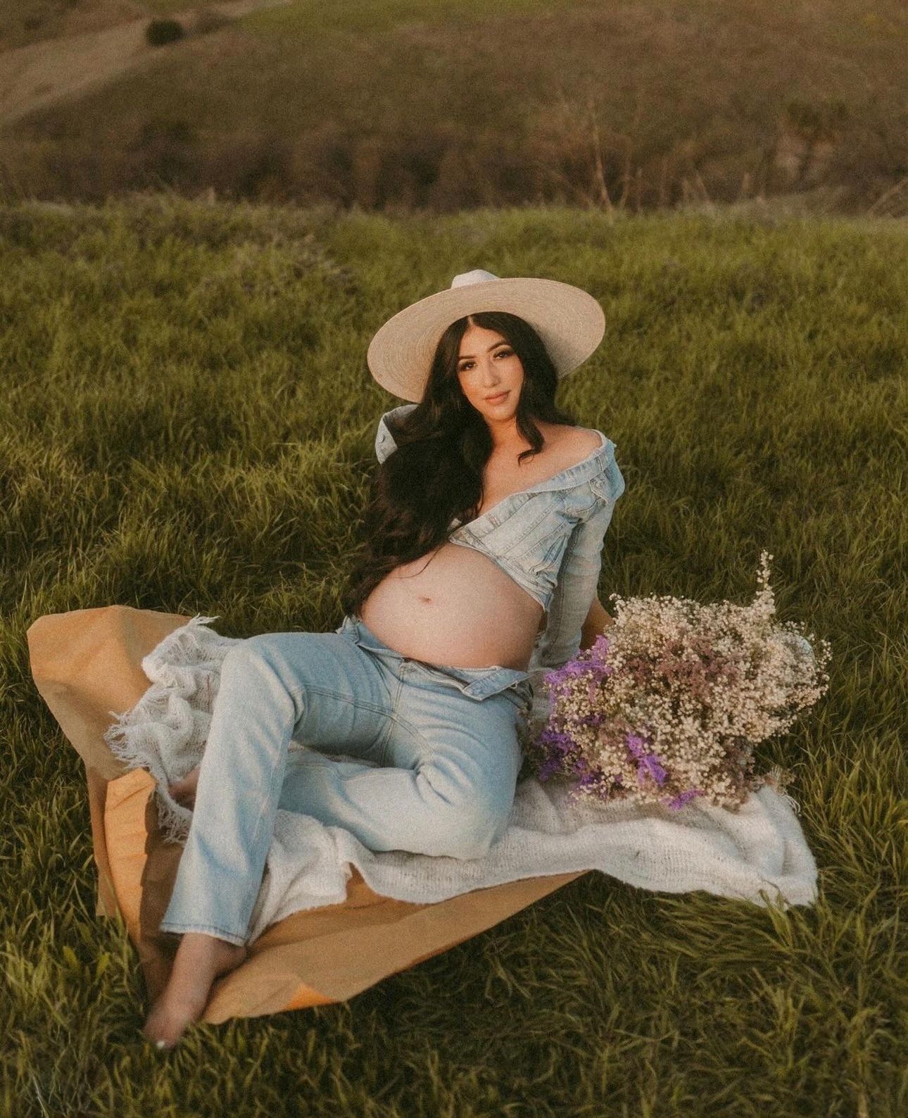Pregnant woman in denim outfit and wide-brim hat sits on blanket in green field holding wildflowers.