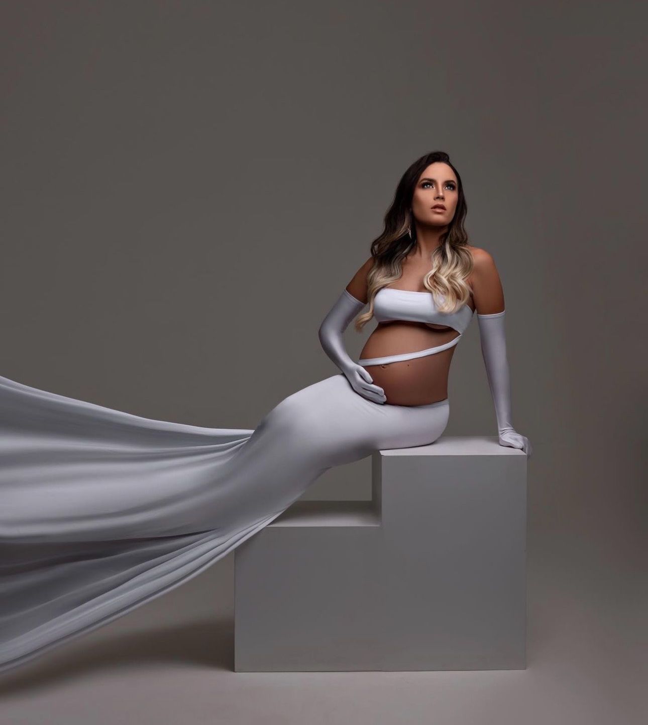 Pregnant woman in elegant white off-shoulder crop top and flowing skirt poses gracefully on white geometric steps.