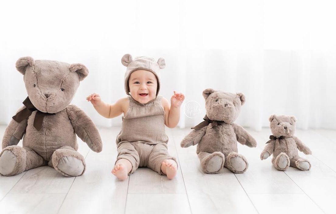 Baby in bear outfit sitting on floor with three teddy bears arranged by size.