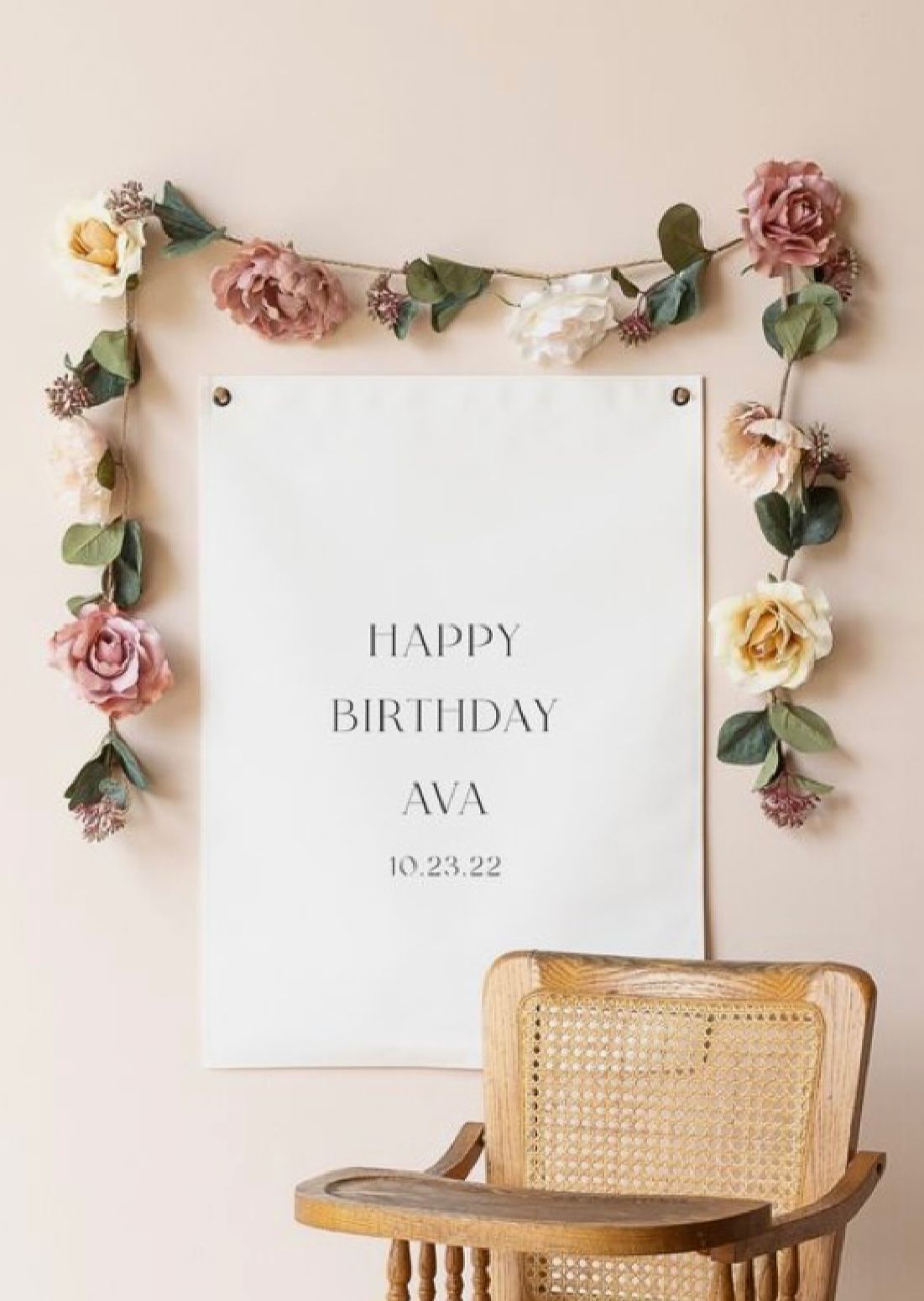 A vintage high chair sits beneath a white birthday sign decorated with delicate pink and yellow roses and greenery.