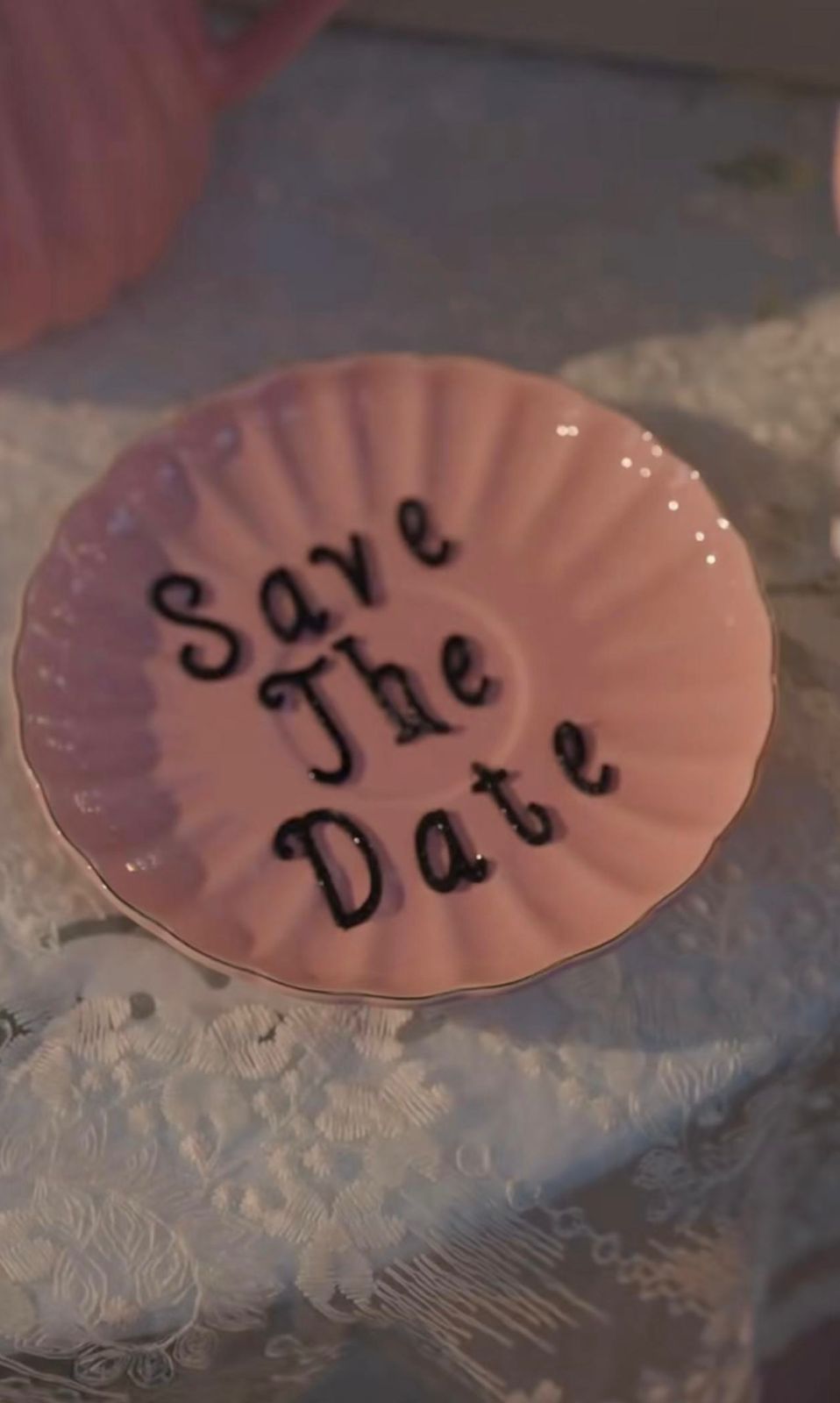 Pink ceramic plate with 'Save The Date' written in black script lettering on a lace tablecloth.