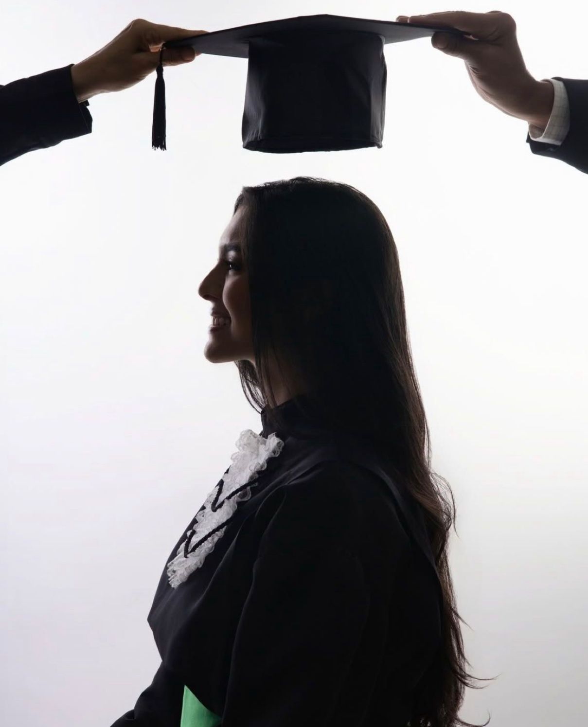 Silhouette of graduate in profile view with hands holding mortarboard cap above in dramatic lighting against white background.