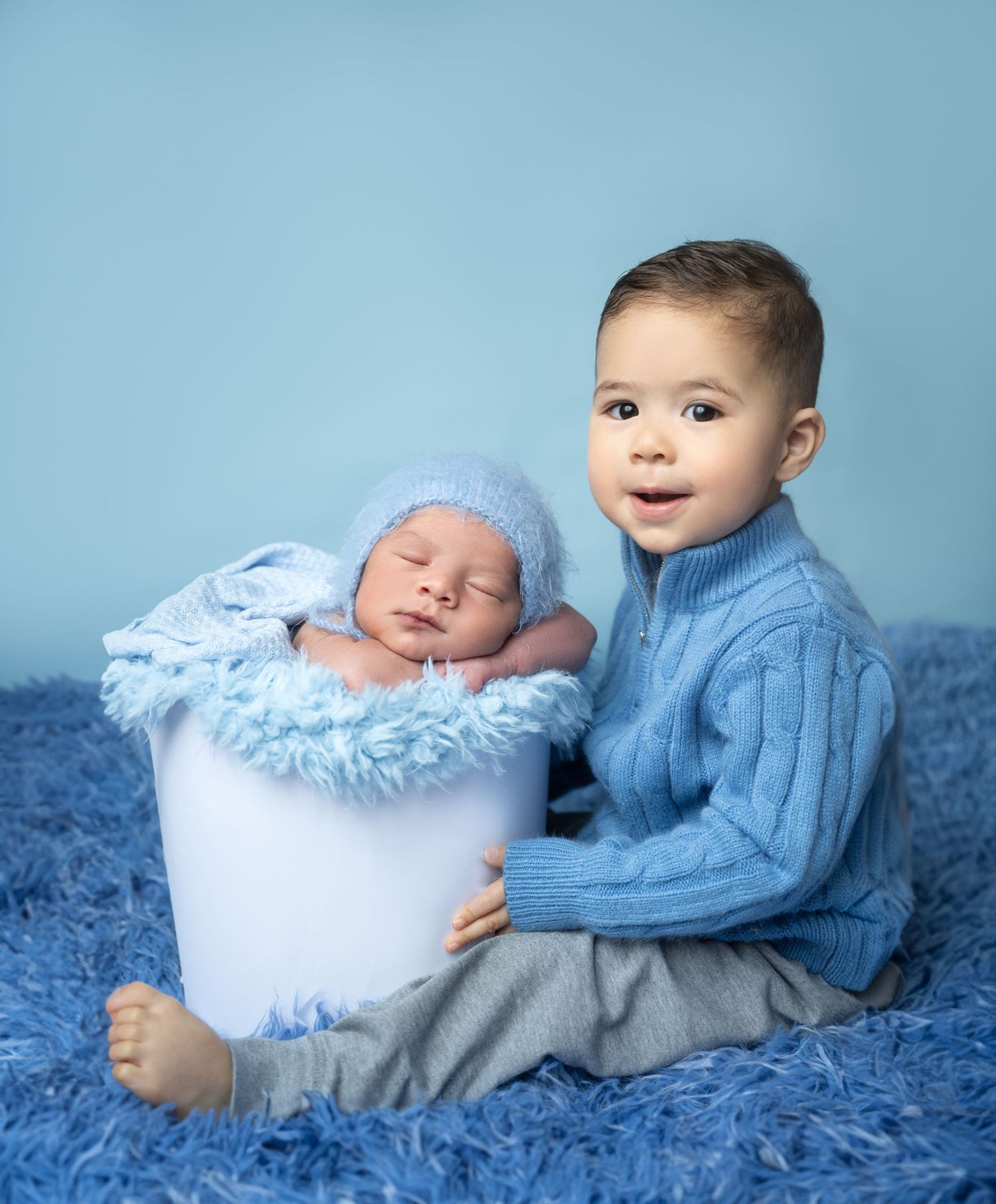 A toddler boy in a blue sweater sits beside a white bucket holding a sleeping newborn baby in a blue hat on a blue fur rug.