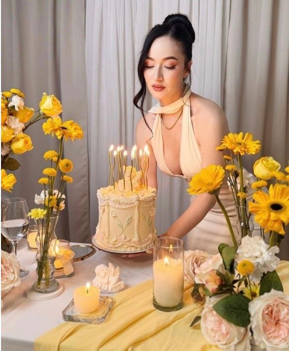 Woman in yellow dress blowing out candles on birthday cake surrounded by yellow flowers and candles.