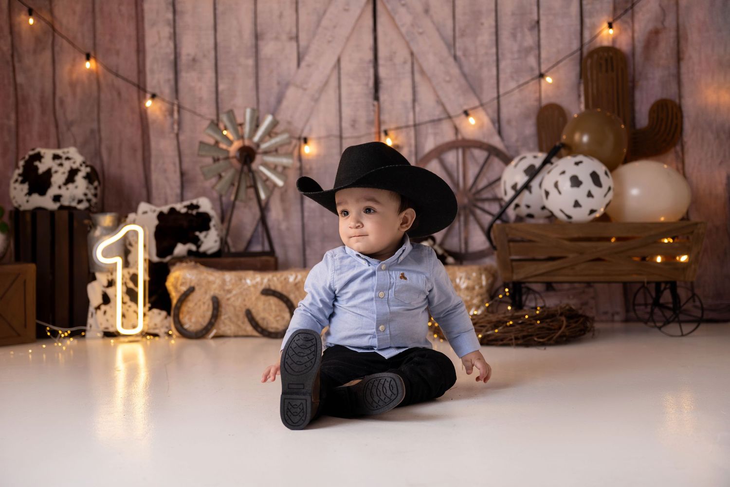 Baby boy in black cowboy hat sits on white floor with western-themed birthday decorations and glowing number one.