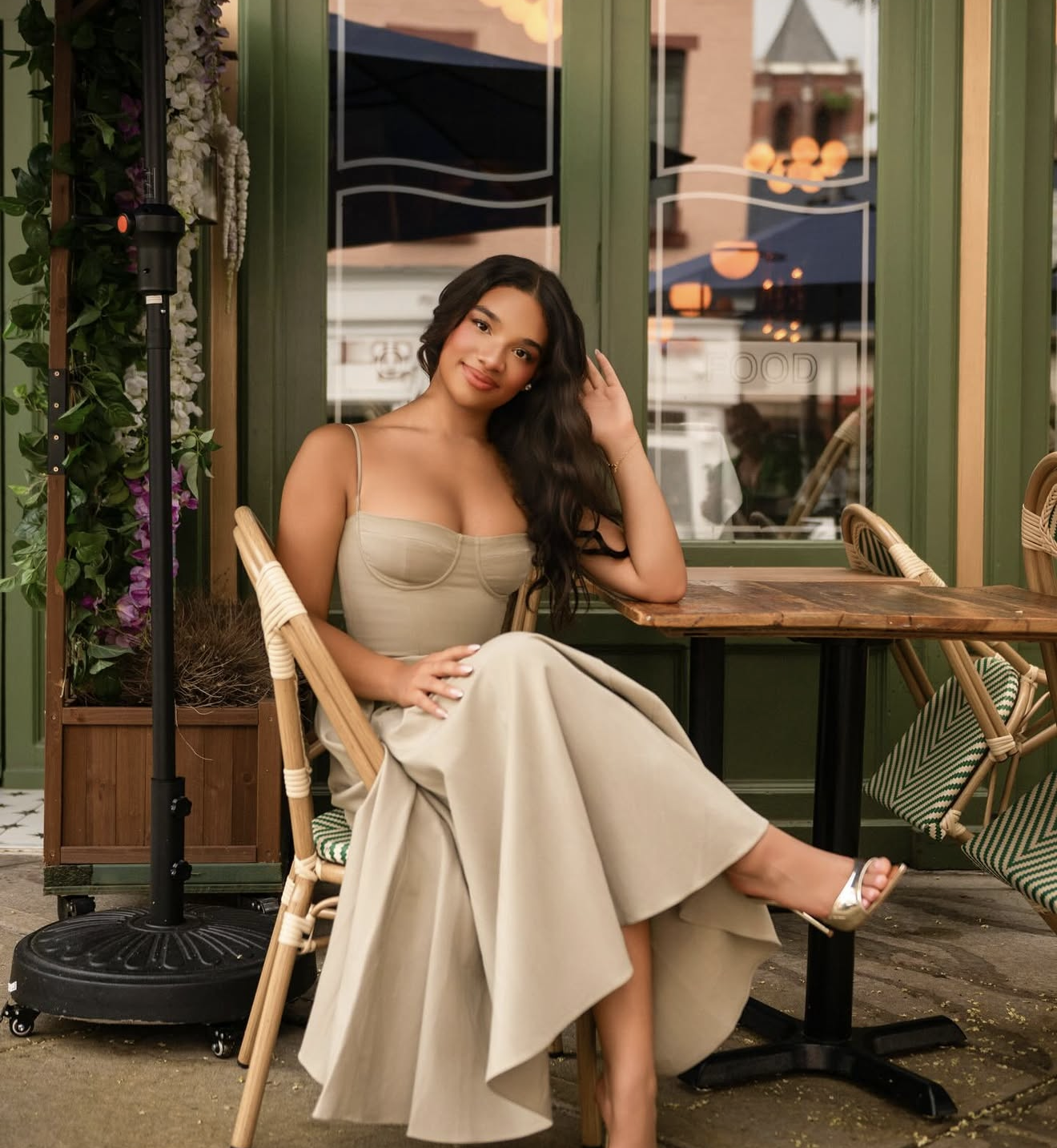 Woman in beige corset top and wide-leg pants seated at outdoor cafe table, posing elegantly in strappy heels.