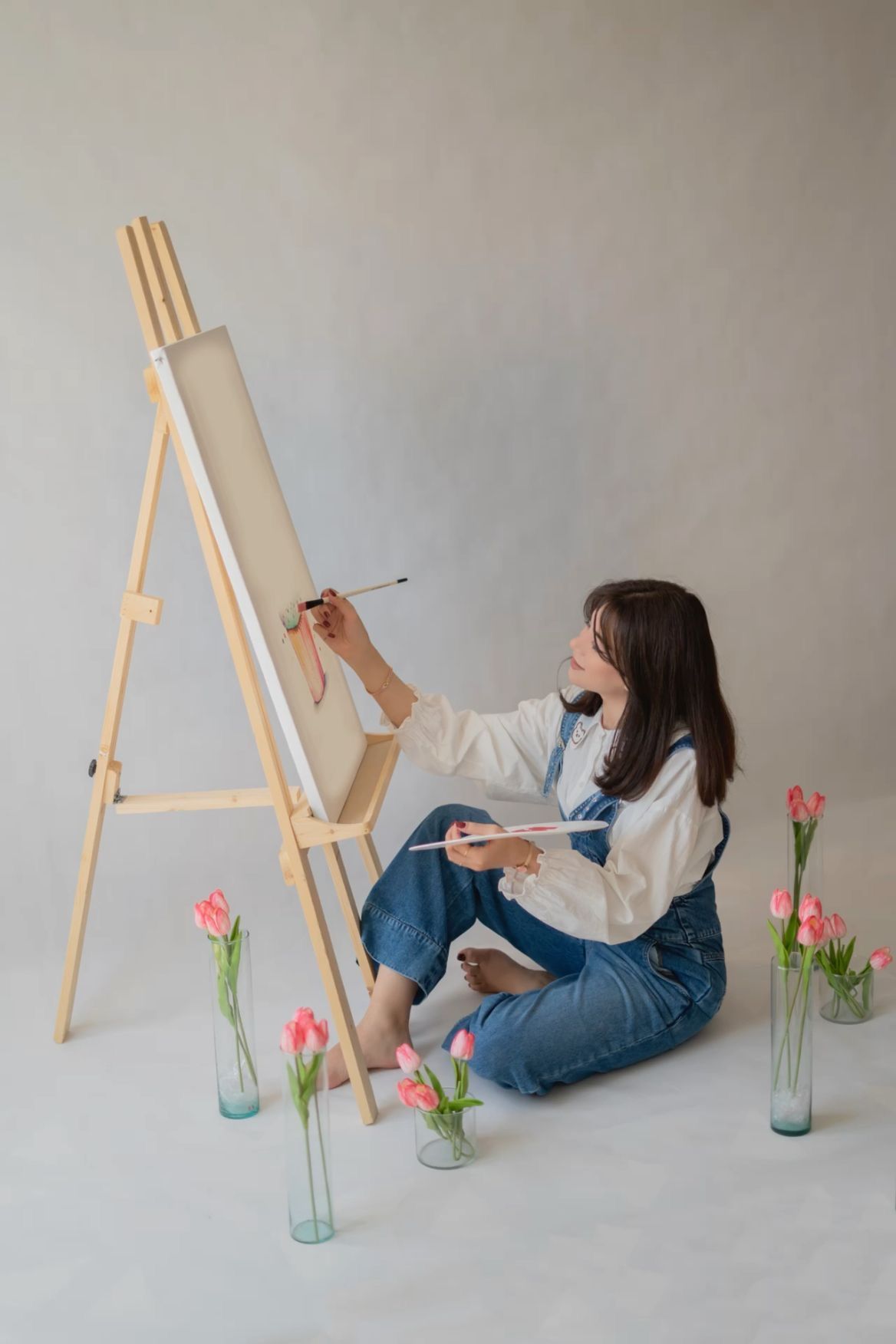 Young woman sitting on floor painting on easel in studio, surrounded by pink tulips in glass vases.