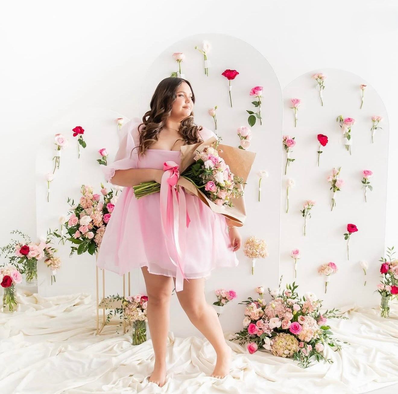 Woman in pink dress holding bouquet surrounded by floral wall decor and arrangements in a bright white studio.