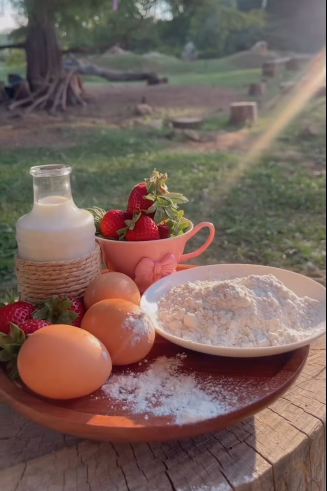 Fresh baking ingredients including eggs, milk, flour, and strawberries arranged on a wooden tray outdoors.
