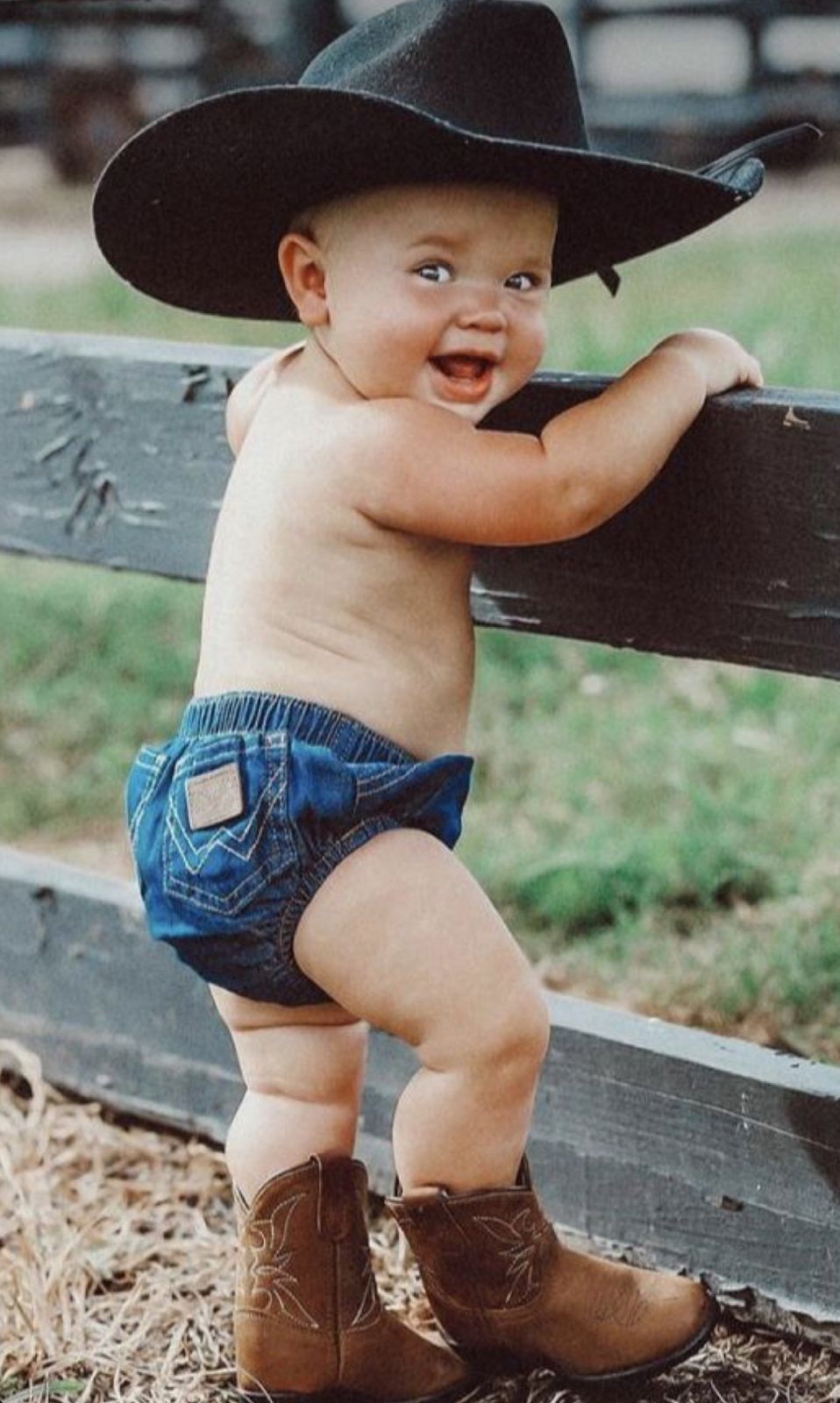 Adorable baby wearing a black cowboy hat, denim diaper cover, and brown cowboy boots, standing at a wooden fence.
