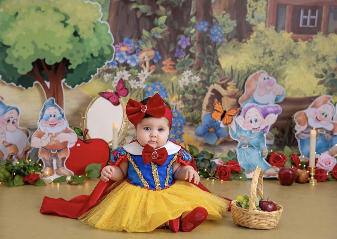 Baby girl in Snow White costume poses with Seven Dwarfs backdrop and red apple for fairy tale themed photo shoot.