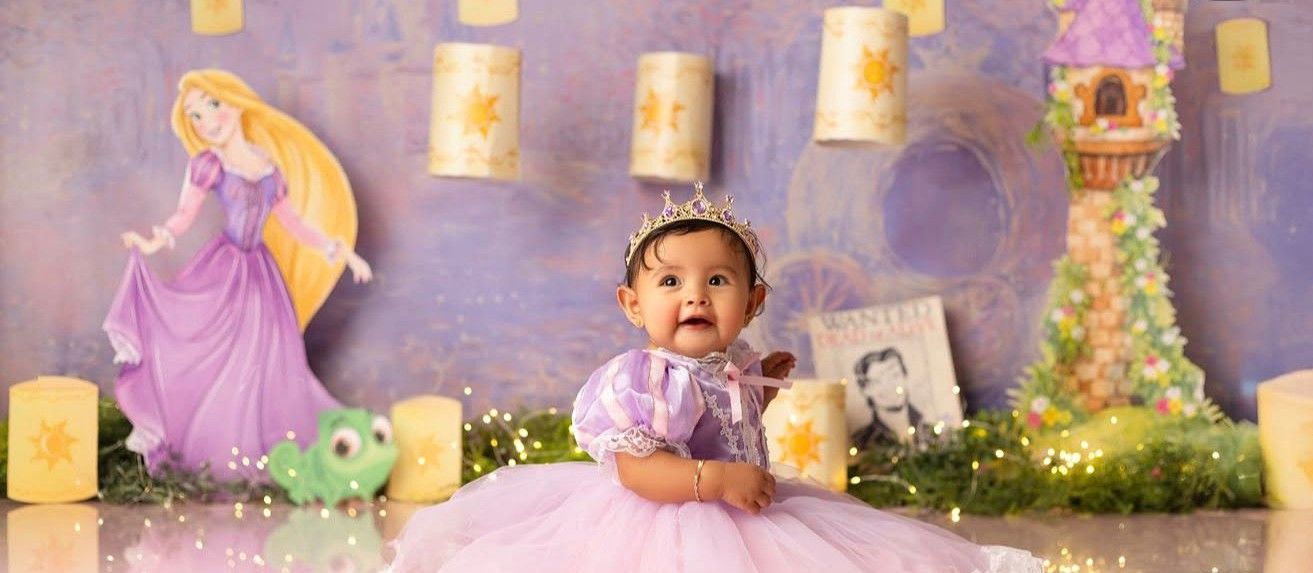 Baby girl in purple tutu sits before a Rapunzel-themed backdrop with tower, lanterns, and fairy lights.