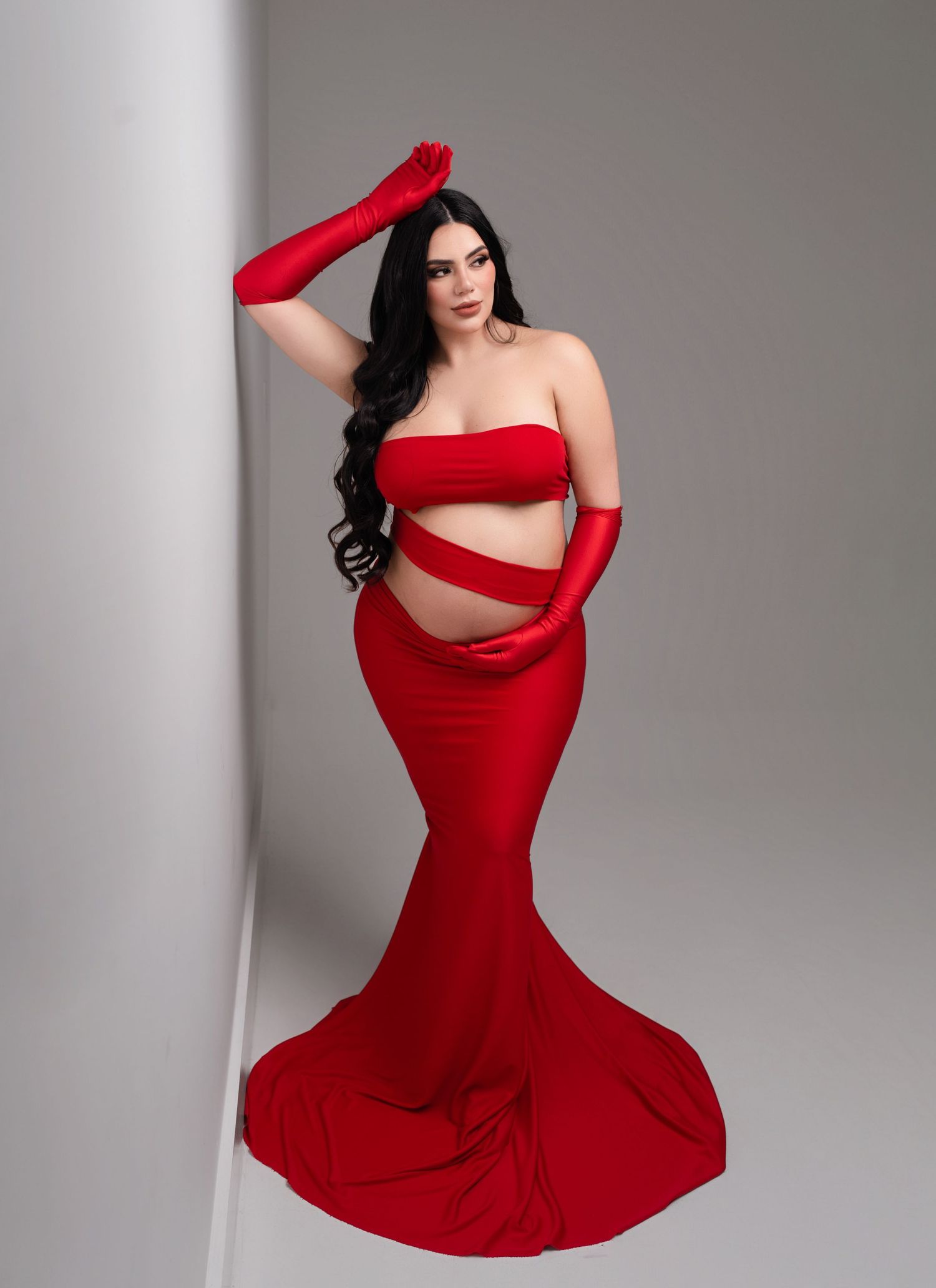 Pregnant woman in red strapless crop top, mermaid skirt, and long gloves posing elegantly in a studio.