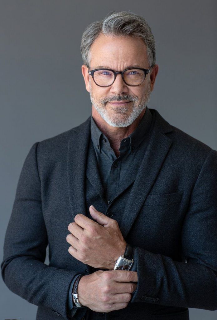 Distinguished middle-aged man with gray hair and glasses wearing a dark blazer, posing confidently against a gray background.