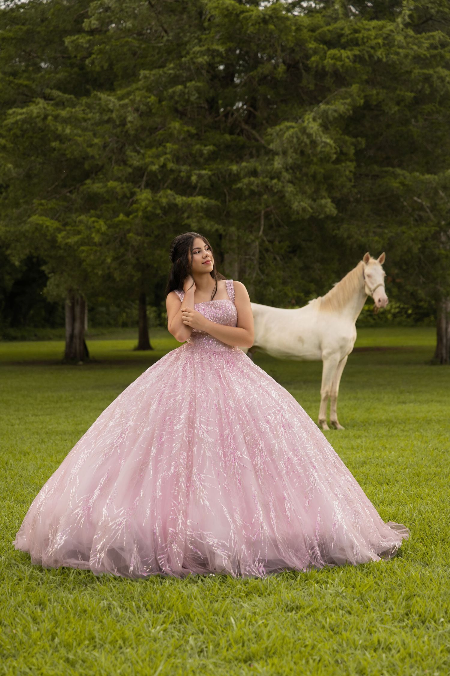 Young woman in pink ballgown quinceañera dress poses with white horse in lush green park setting.
