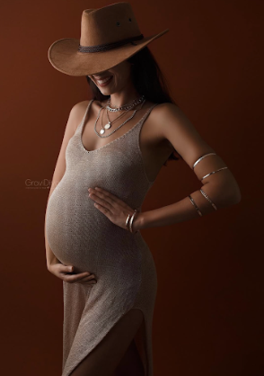 Artistic maternity portrait featuring a pregnant figure in a gray dress and brown cowboy hat against a brown backdrop.