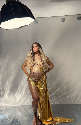 Model poses dramatically in a flowing gold dress against a grey studio backdrop.