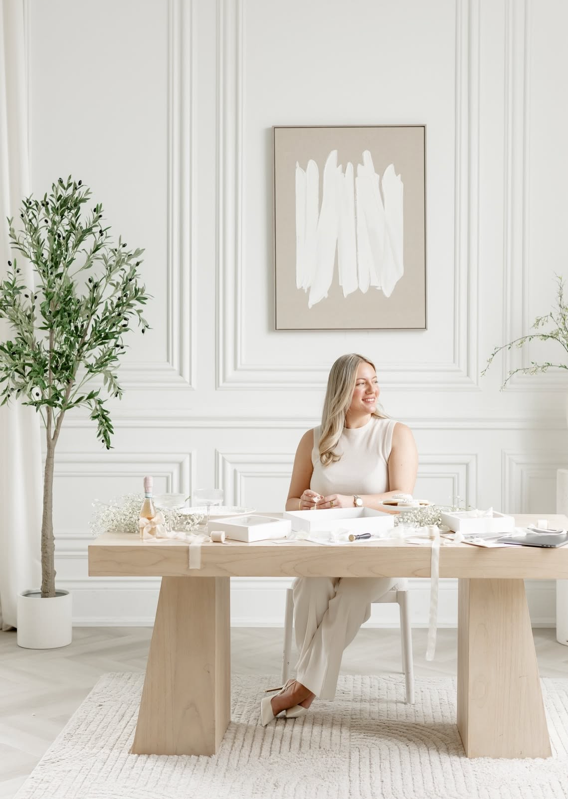 Woman smiling at a light wood desk in a bright white office with abstract art and olive trees.