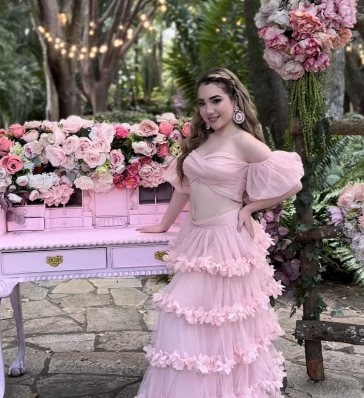 Young woman in a pink off-shoulder tiered gown posing beside a pink piano surrounded by floral arrangements outdoors.