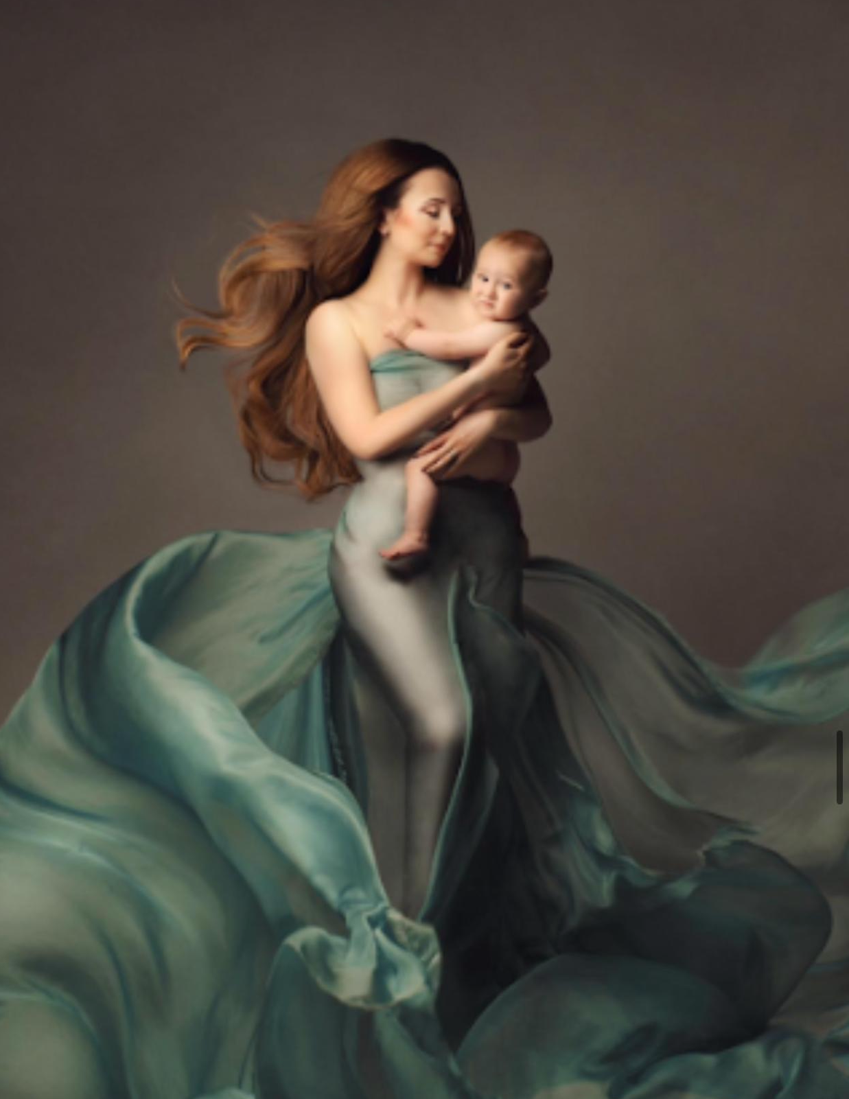 A mother in flowing teal fabric holds her baby, her long hair flowing gracefully in a studio portrait.