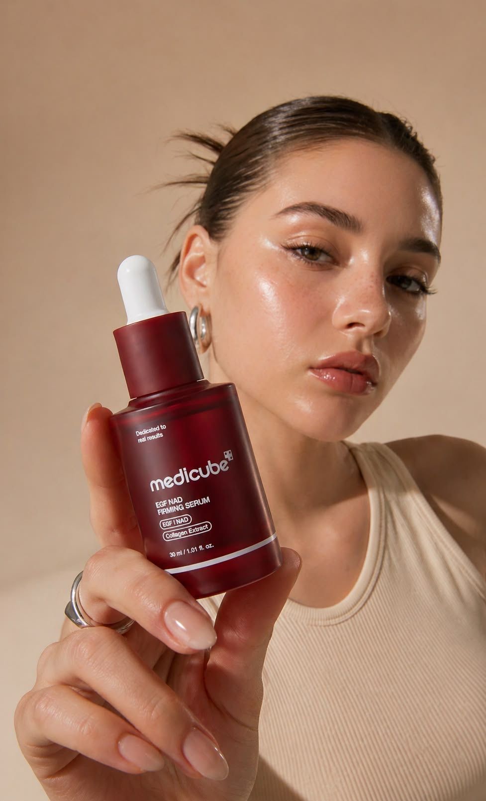 Young woman holding a Medicube EGF NAC Firming Serum bottle, showcasing glowing skin against a neutral background.