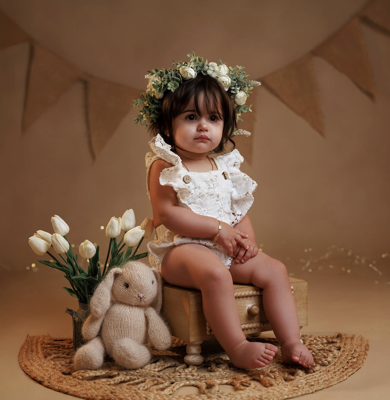 Toddler girl in white lace romper and floral crown sits on wooden box with bunny toy and tulips.