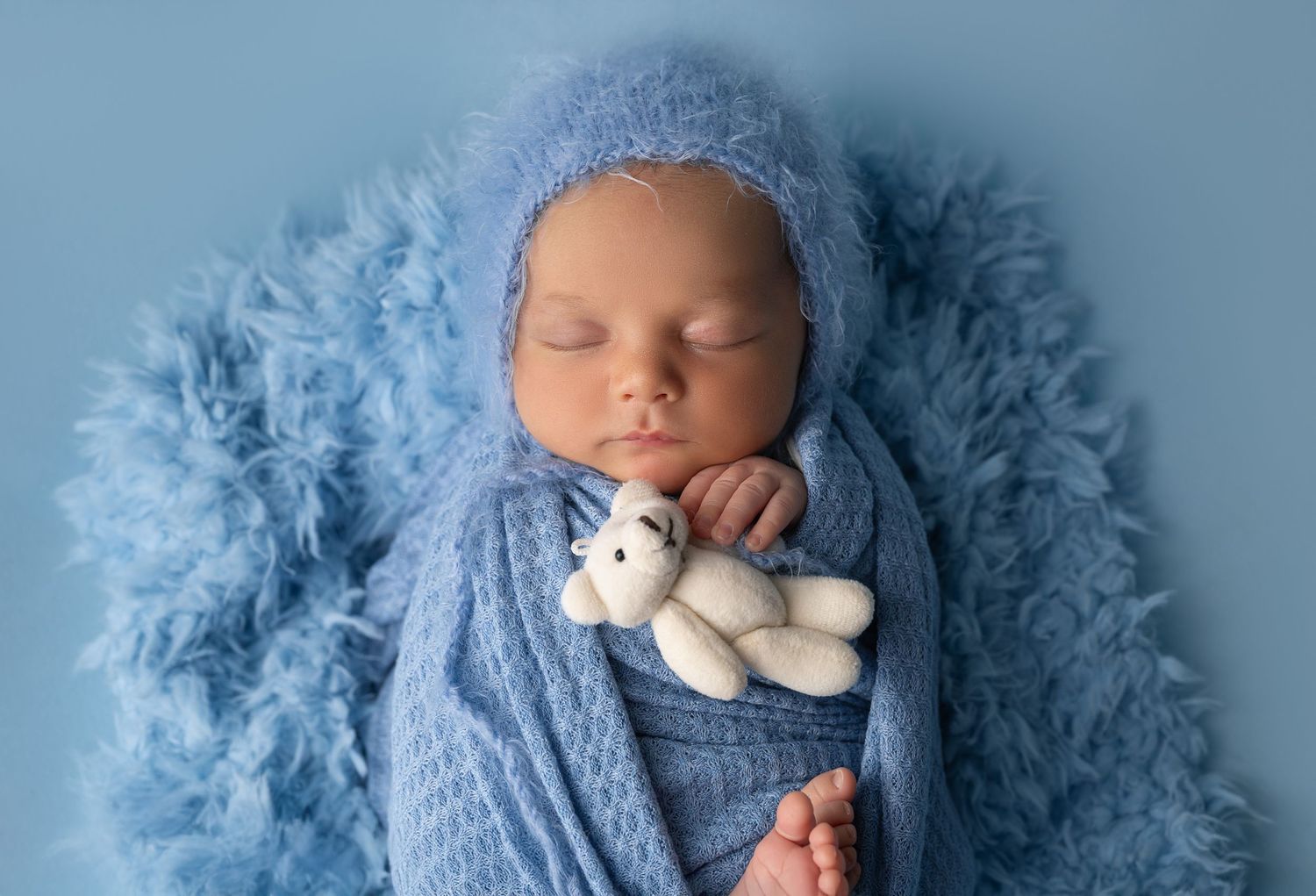 Cozy newborn wrapped in blue knit holding tiny teddy bear on fluffy backdrop.